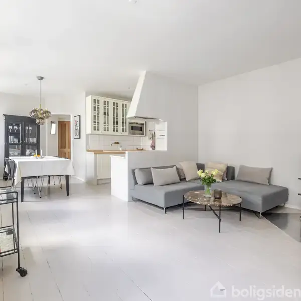 En stue med en grå sofa og sofabord, stående ved en hvid væg; i baggrunden ses et spisebord og et åbent køkken med skab.
