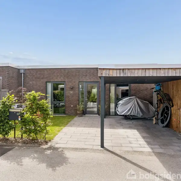 En moderne, lav bolig med fladt tag i mursten. Foran huset står en overdækket carport med en motorcykel dækket af et gråt tæppe. Omkring er der grøn beplantning og flisebelægning.