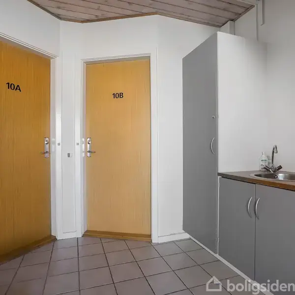 To døre, 10A og 10B, står i et lille gangareal. En grå skabsenhed med en vask er placeret til højre. Flisegulv og hvidmalede vægge.