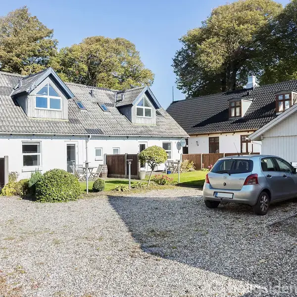 To biler holder parkeret på en grusopkørsel foran et hvidt parcelhus med kviste og skråtag. Omkringliggende huse og træer står i baggrunden, skaber en boligområdeatmosfære.