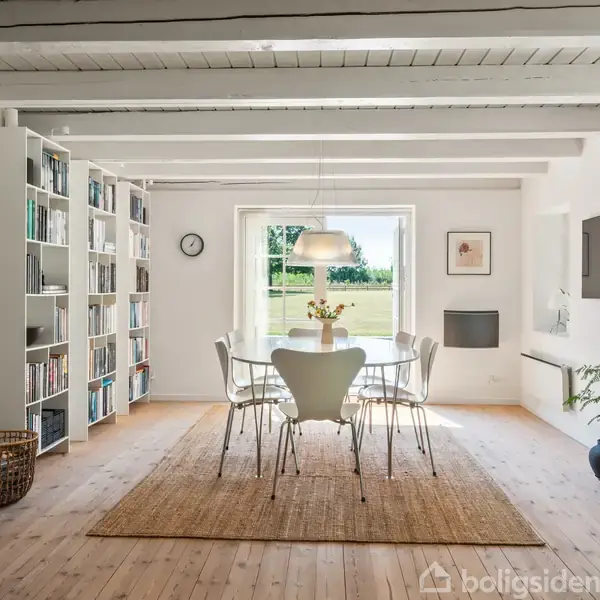 Spisebord med seks stole står på tæppe, under lampe i lyse, moderne stue. Reol fyldt med bøger til venstre, stor vinduesudsig til have foran. Potteplante og vægur i rummet.