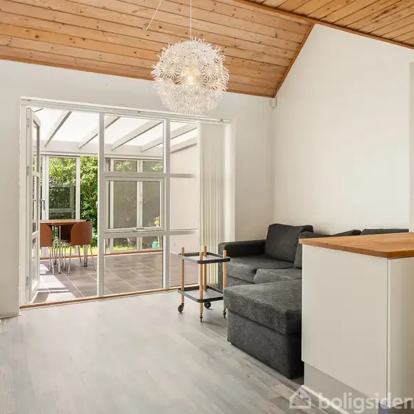 Stue med hjørnesofa mod vinduesparti, der fører til overdækket terrasse med udsigt til grøn have. Træloft og hængende lampe. En lille reol står i hjørnet.