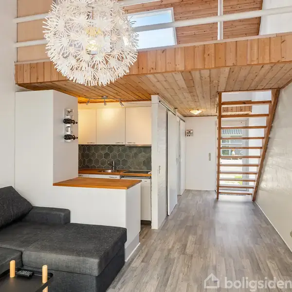 En moderne stue med en mørkegrå sofa til venstre, en åben loftsænket trappe til højre, og et kompakt, hvidt køkken med træbordplade i midten. En dekorativ loftlampe hænger over stuen.