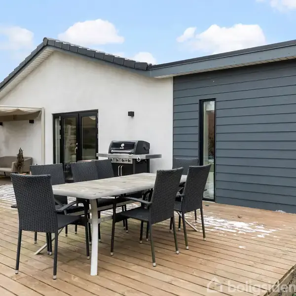 Et moderne hus med en træterrasse har et sæt havemøbler med seks stole. Der står også en grill på terrassen. Huset har grå og hvide vægge under en blå himmel.