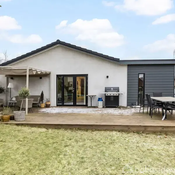 Hus med moderne design står på en terrasse. Havemøbler og grill står klar, omgivet af en stor, velplejet græsplæne. Skyfri blå himmel danner baggrund.