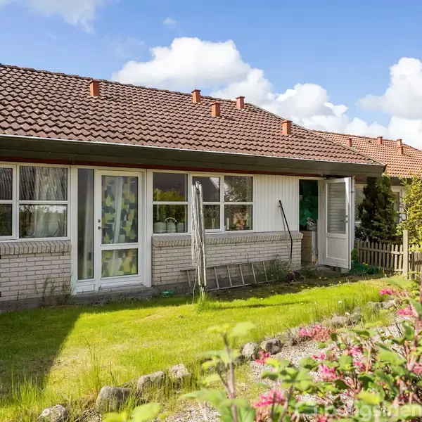 En rækkehus med rødt tegltag og store vinduer. Havedør åben til grøn forhave med blomster. Sollys og blå himmel i baggrunden.