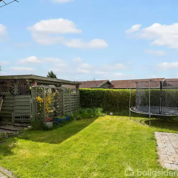 Trampolin står i en grønt have med hæk omkring. Ved siden af er der et skur og nogle blomster. Himlen er blå med skyer.