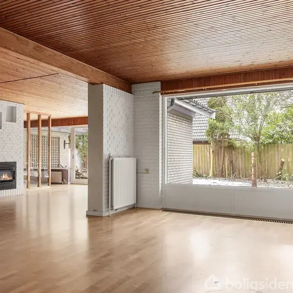 Et rummeligt, lyst stueområde med trægulv og træloft. En moderne brændeovn er tændt i hjørnet. Store vinduer viser en udsigt til en have med træer og hegn.