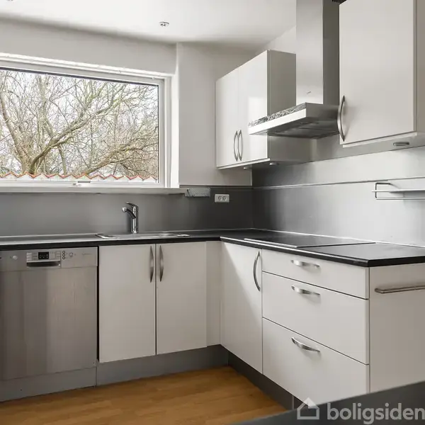Moderne køkken med hvide skabe og stålgreb. Opvaskemaskine ved siden af vasken under et vindue. Grå backsplash, trægulv og en stor emhætte. Udsigt til udenfor.