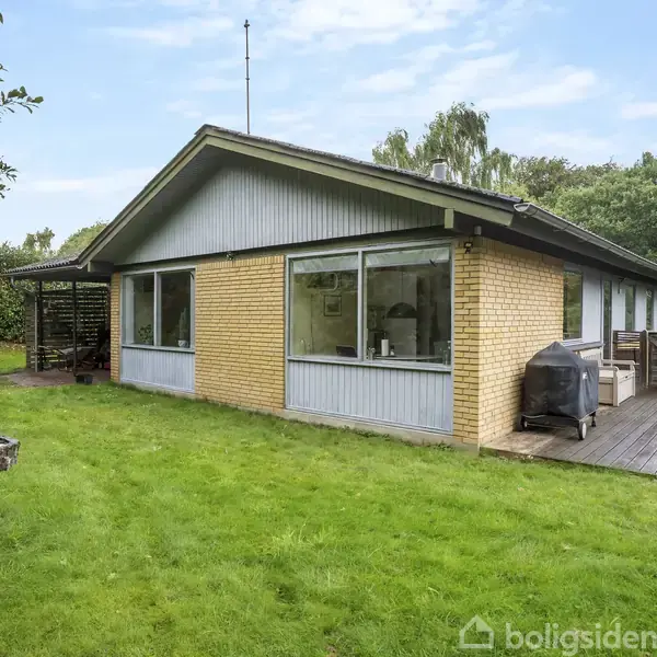 Et etplanshus med gule mursten og store vinduer står i en have med grøn græsplæne. Der er en terrasse med grill, og træer omgiver området.