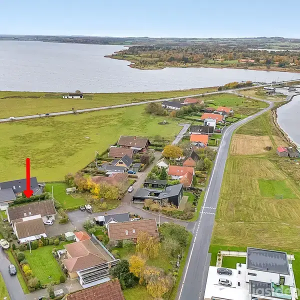 Hus markeret med en rød pil ligger i et boligkvarter nær kysten. Huset er omgivet af andre boliger og grønne arealer, med en stor vandflade i baggrunden.