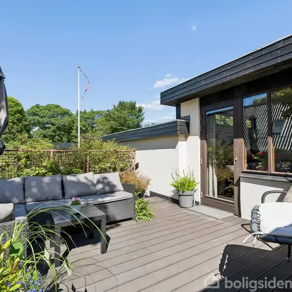 Terrasse med sofa- og loungemøbler i solrig gårdhave, omgivet af planter og træer. Flagstang kan ses i baggrunden. Solrig dag med blå himmel og hvide skyer.