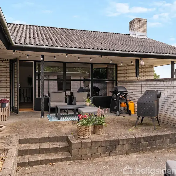 Terrasse med udendørs møbler og grill. Sofa og bord i midten. Lav murstensvæg omgiver området. Planter i potter tilføjer grønt. Bygningens vinduer i baggrunden.