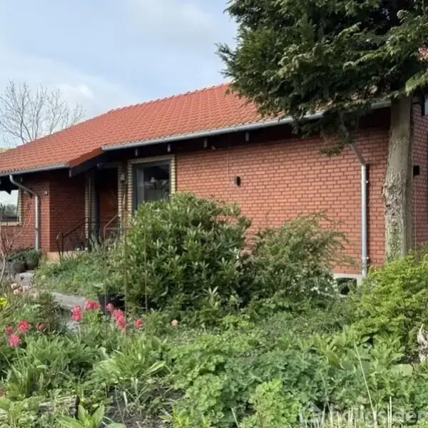 Rød murstenshus med rødt tegltag, omgivet af en frodig have med blomster og buske. Et dansk flag vajer i baggrunden, og en stor træ står tæt på facaden.