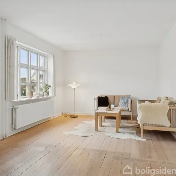 En stue med en sofa og sofabord står på et lyst trægulv. Rummet har hvide vægge, en lampe i hjørnet, og vinduer til venstre, der lukker lys ind.