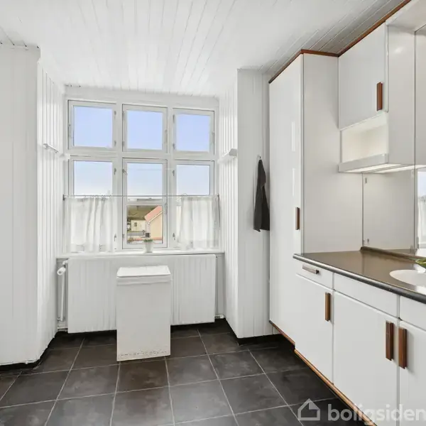 Et lyst køkken med hvide skabe har et sort flisegulv. Vinduer langs bagvæggen giver udsigt over hustage og skyfri himmel. En vask med hvid kande står på køkkenbordet.