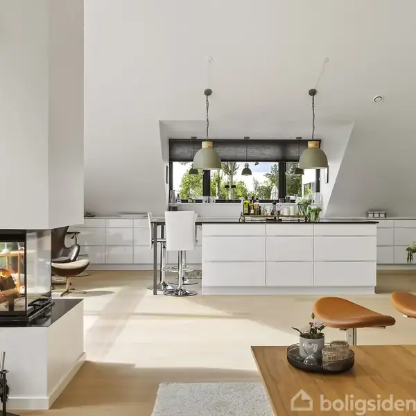 Pejs med ild står til venstre i en moderne, lys stue. I baggrunden er et åbent køkken med hvide skabe, barstole og hængelamper. Grønne planter tilføjer liv til rummet.