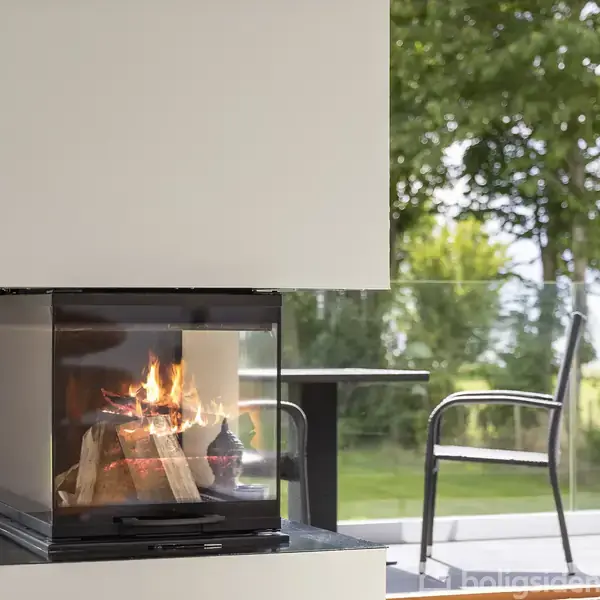 Pejs med brændende træ, står i en moderne stue med store glasdøre. Udenfor ses terrasse med stole og bord, omgivet af frodig natur.