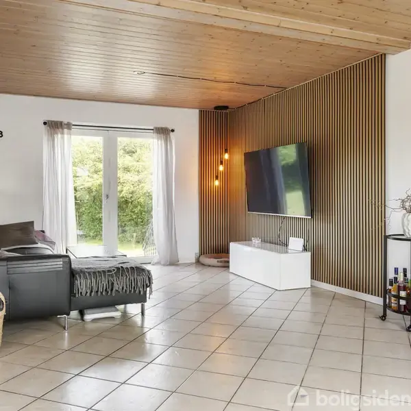 En stue med sort sofa står mod en hvid væg med et stort vægur. Et fladskærms-TV hænger på en trælamelvæg. Store vinduer med gardiner slipper lys ind. En kurv med puder står ved siden af sofaen.