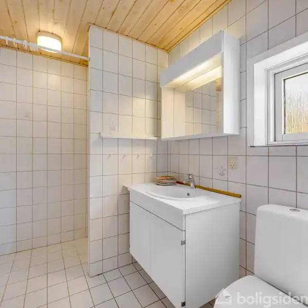 En hvidt flisebeklædt badeværelse med et toilet og et hvidt skab med vask. Der er en oplyst spejlskab over vasken og et lille vindue til højre. Træloft med loftslampe.