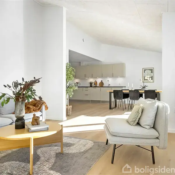 Moderne stue med lyse møbler og sofabord, dekoreret med planter og puder, oplyst af naturligt lys. I baggrunden ses et åbent køkkenområde med spiseplads og indretning i neutrale nuancer.