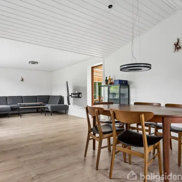 Stue med en rund træspisebord og fire stole i forgrunden, grå sofa i hjørnet, lyse vægge og trægulv. En fladskærm hænger på væggen, med vinduer i baggrunden.