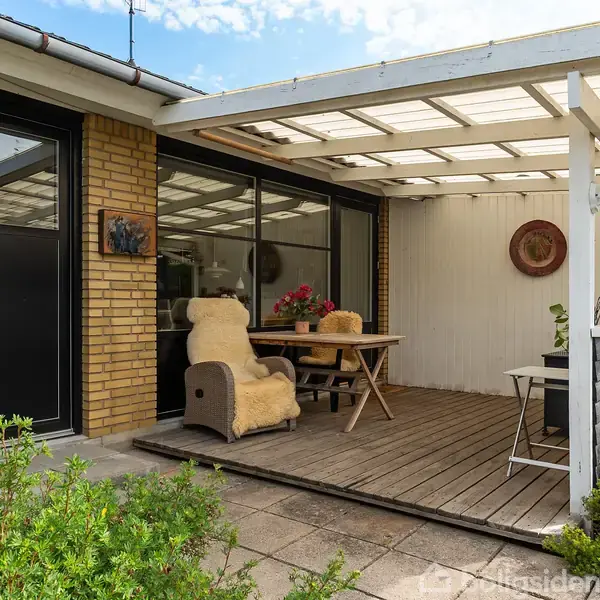 Terrasse med to komfortable stole og et bord er placeret under et halvtag ved siden af en gulstensvæg. Planter omgiver området, og der er en dør med tallet "17" på væggen.