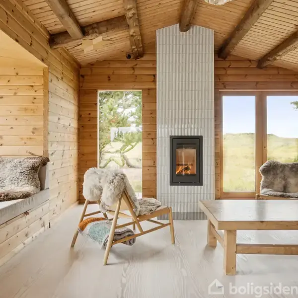 En hyggelig stue med trævægge og loftbjælker, har en lit pejs som midtpunkt, omgivet af stol og sofa med pels. Vinduer viser en udsigt til naturen udenfor.