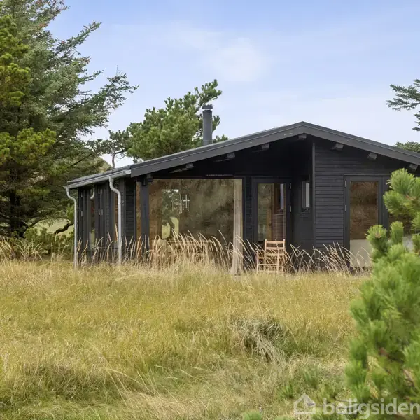 Sort sommerhus står på en græsmark omgivet af træer under en blå himmel. Vinduer reflekterer omgivelserne og en enkelt stol ses ved indgangen.