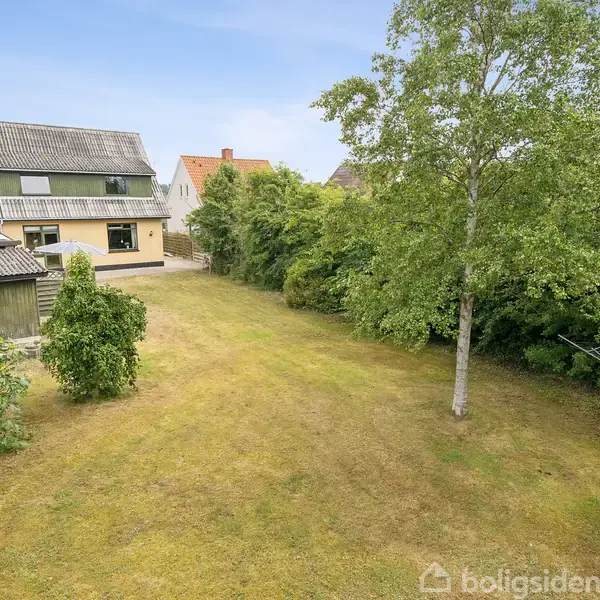 Hus set fra haven; grønt græs og store træer i forgrunden; flere boliger bagved; klar blå himmel i baggrunden.