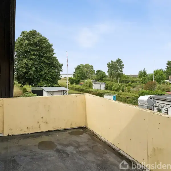 En åben altan med gule vægge. Udsigten viser en have med grønne træer og buske samt nogle huse. Himlen er overskyet med blå nuancer.