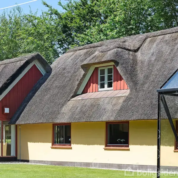 Stråtækt hus med rødt bindingsværk og gule vægge. Det har små vinduer og ligger i en grøn have omgivet af høje træer.