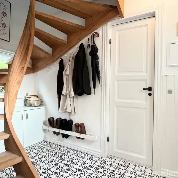 En trævindeltrappe leder opad i en entré med mønstrede sorte og hvide fliser. Tøj og sko hænger og står tæt ved en hvid dør, der åbner ind til et rum.