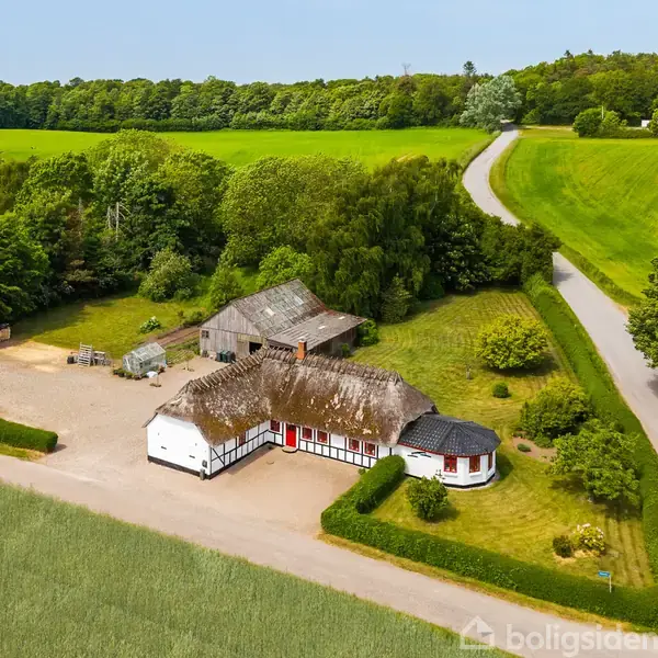 En gård omgivet af marker og skov ligger ved en snoet vej. Der ses flere bygninger med stråtage, omkranset af grønt landskab og udsigt til vand i baggrunden.