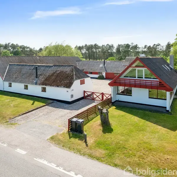 Gårdbygning med stråtag og hvid facade står stille ved en vej. Omkring bygningen er der græsplæner og skov i baggrunden. En terrasse med rødmalet rækværk er synlig.