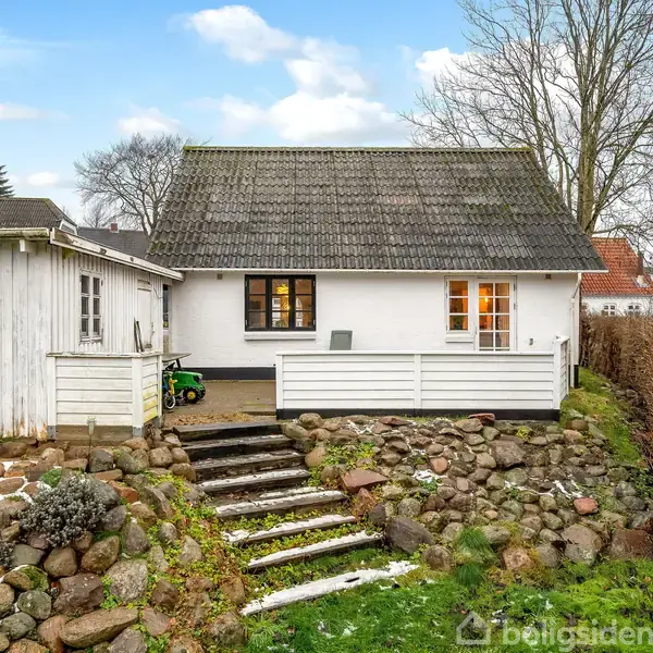 Et hvidt hus med sort tag står fredeligt. Trappetrin fører op til indgangen. Omgivet af stenvægge og en frodig have under en klar blå himmel.