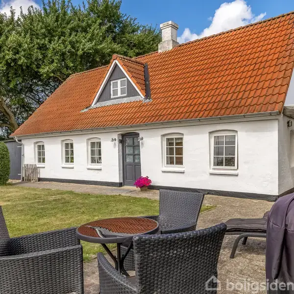 Hvidt hus med rødt tegltag står stille i en grøn have. Foran huset er et rundt havebord med fire stole. Blå himmel og træer omkranser ejendommen.