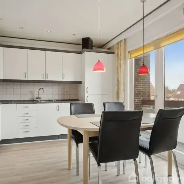 Et moderne køkken med hvide skabe og en indbygget ovn. Fire sorte stole omkranser et rundt bord. To røde lamper hænger over bordet. Store vinduer giver udsigt udenfor.