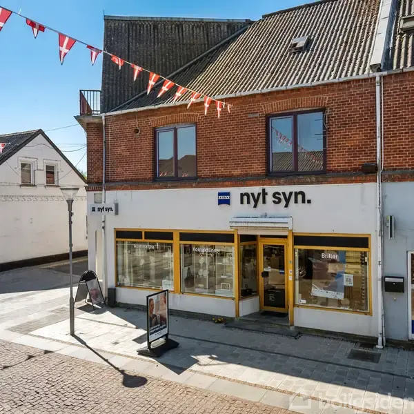Butik med skiltet "nyt syn." og store, gule vinduer. Facaden er af rød mursten. Omgivelserne har brostensbelægning, flagrækker og nabolokale med skilt "matas" til højre.