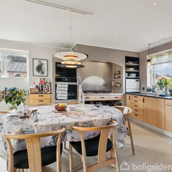 Et køkken med et rundt spisebord dækket af en dug med blomster. Bordet er omgivet af fire træstole. Der er moderne træskabe, en ovn og store vinduer med udsigt til en have.