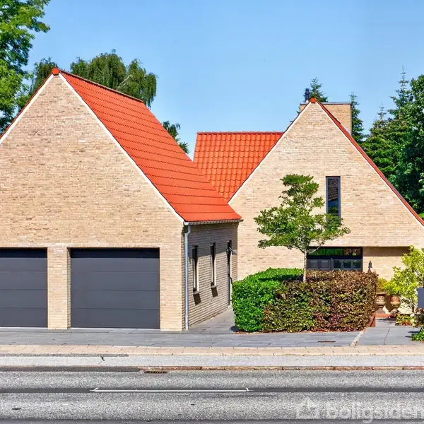 Et moderne hus med rødt tegltag og lyse mursten. To store, sorte garageporte dominerer forgrunden, omgivet af hække og træer. Beliggende på en fortovskantet vej med grønne omgivelser.