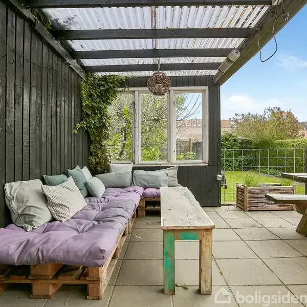 Sofa af træpaller med farverige puder står under overdækket terrasse med trævægge. Et træbord og bænke er placeret ved siden, med udsigt til en grøn have med træer og buske.