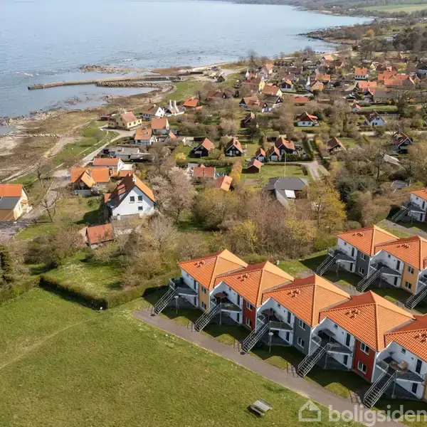 Hus med gråtag ligger ved havet i en landlig setting. Foran huset er grønne buske og træer. Havet strækker sig til horisonten, der er en klar blå himmel.