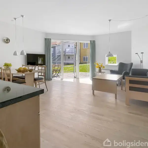 Lys stue med spisestuemøbler og sofa, store vinduer åbner ud til en have. Udendørs kan man se en moderne bygning. Gulvet er lyst træ og rummet har minimalistisk indretning.