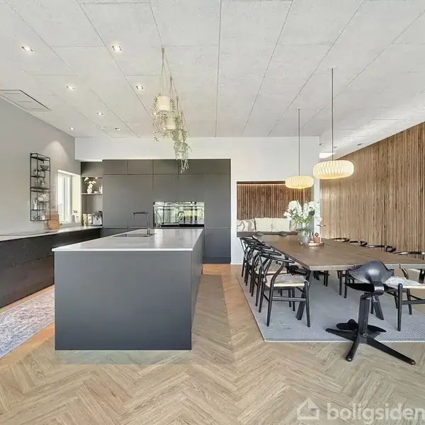Et moderne køkken med antracitgrå skabe og en stor ø opstillet. Ved siden af er der en spisestue med et langt bord og sorte stole. Trævægge og hængelamper skaber en elegant atmosfære.