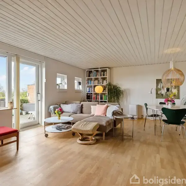 En stue med sofa, sofabord, og reoler. Et spisebord med stole står i baggrunden. Store vinduer giver udsyn til en terrasse med planter. Lys træparketgulv og hvide vægge.
