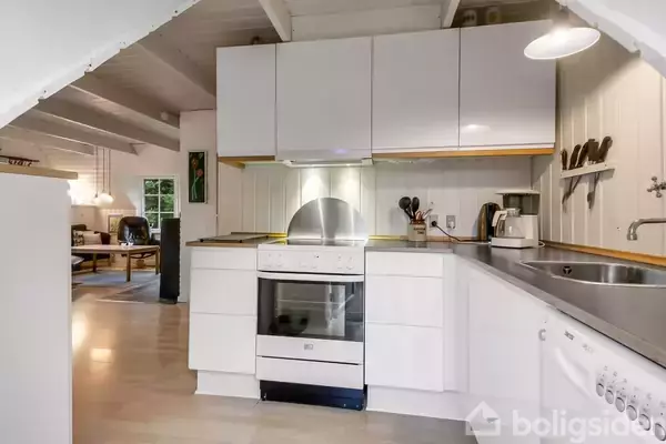 Et hvidt køkken med en komfur, emhætte og opvaskemaskine i et hjemligt rum. Køkkenet er lyst op med hængende lamper, og i baggrunden ses en stue med møbler.