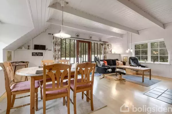 Et lyst opholdsrum med spisebord, stole og sofagruppe. Store vinduer og glasdør giver udsigt til skov. Rummets lyse trævægge og lofter skaber en åben og indbydende atmosfære.