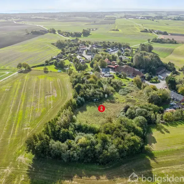 En stor græsmark omgivet af træer, markeret med et rødt ikon "B," ligger i kanten af en landsby med spredte huse og åbne marker i det fjerne.