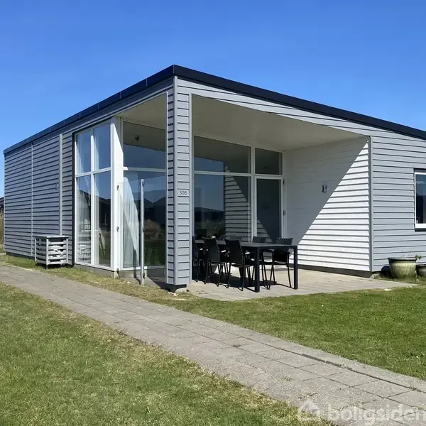 Et moderne, rektangulært sommerhus med lysegrå træbeklædning har store vinduer. En overdækket terrasse med sort bord og stole står foran. Huset er omgivet af en græsplæne og flisegang.